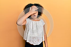 Young little girl with afro hair wearing casual clothes peeking in shock covering face and eyes with hand, looking through fingers