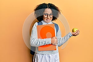 Young little girl with afro hair wearing school bag holding books and green apple clueless and confused expression