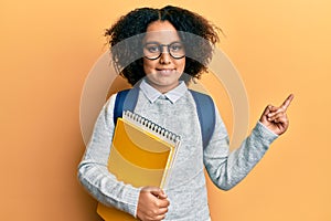 Young little girl with afro hair wearing school bag and holding books smiling happy pointing with hand and finger to the side