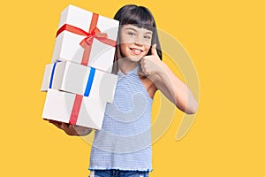 Young little girl with bang holding gifts smiling happy and positive, thumb up doing excellent and approval sign