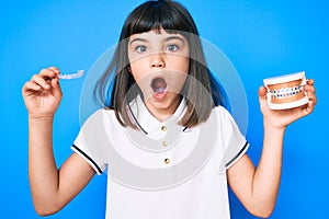 Young little girl with bang holding invisible aligner orthodontic and braces afraid and shocked with surprise and amazed