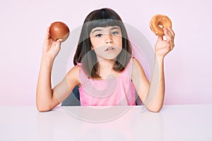 Young little girl with bang holding red apple and donut sitting on the table clueless and confused expression