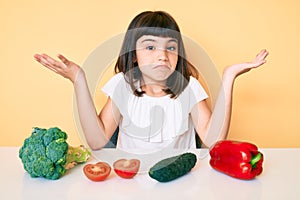 Young little girl with bang sitting on the table with veggies clueless and confused with open arms, no idea and doubtful face