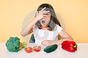 Young little girl with bang sitting on the table with veggies peeking in shock covering face and eyes with hand, looking through