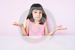 Young little girl with bang wearing casual clothes sitting on the table clueless and confused expression with arms and hands