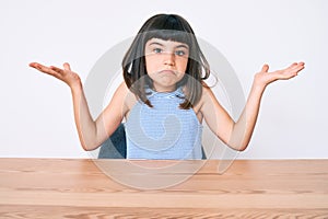 Young little girl with bang wearing casual clothes sitting on the table clueless and confused with open arms, no idea and doubtful