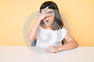 Young little girl with bang wearing casual clothes sitting on the table peeking in shock covering face and eyes with hand, looking