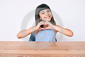 Young little girl with bang wearing casual clothes sitting on the table smiling in love doing heart symbol shape with hands