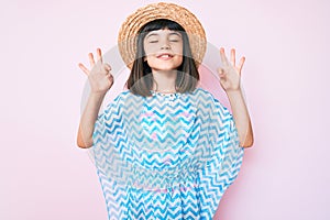 Young little girl with bang wearing summer dress and hat relax and smiling with eyes closed doing meditation gesture with fingers