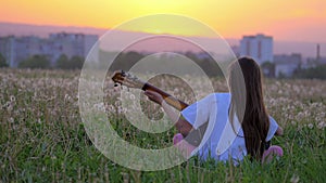 Young little girl plays acoustic guitar. sitting with his back to camera away from home field dandelions at sunset. concept lifest