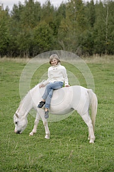 Young little girl in a white sweater and jeans sitting cross-legged on a white horse. Lifestyle portrait