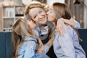 Young little girls child kiss overjoyed middle-aged mom relaxing together on sofa in living room