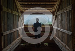 Young lonely teenage boy standing near the wooden forest house window and enjoying the morning spruce foggy forest. Traveling and