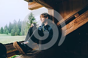 Young lonely teenager boy drinking hot tea from thermo flask while he sitting on the wooden forest house terrace and enjoying the