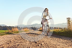 Young long hair woman with sunglasses riding bike on dusty country road, afternoon sun lit country background