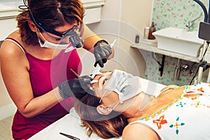 Young long-haired brunette woman putting false eyelashes on lady in beauty salon with masks