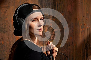 A young long-haired DJ girl in a black top, black baseball cap and headphones poses against a grunge brown wall and