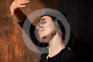 A young long-haired DJ girl in a black top, black baseball cap and headphones poses against a grunge brown wall and