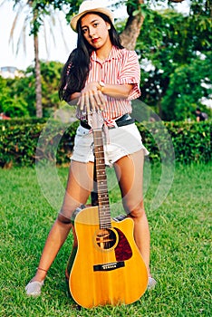 Young long-haired Latin girl, wearing red and white striped shirt and hat, standing on a garden lawn playing an acoustic guitar