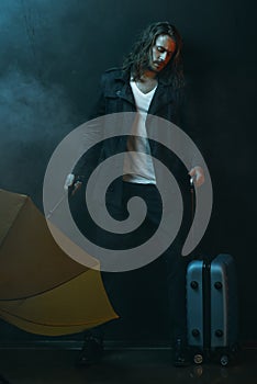 Young long haired man holding yellow umbrella and suitcase