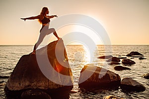 Young long-haired woman in warrior pose from yoga standing on a big stone in water. Sunset, relaxation, meditation