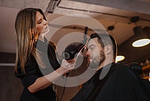 Young long long-haired female hairdresser hairstyling drying hair with professional hair dryer bearded man in Salon Chair. Modern