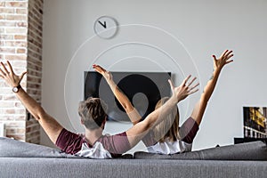 Young in love couple watch tv with raised hands celebrate win sitting on couch