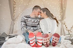 Young lovely couple beautiful wife and handsome husband both wearing warm sweaters and festive socks sitting in bedroom decorated
