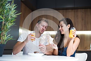 Young lovely couple eating croissant and drinkink orange juice for breakfast in the kitchen playing with bagels start of