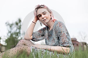 Young lovely woman with pink hair in stylish summer blue dress in black vintage tights sits on fresh green grass outdoors on