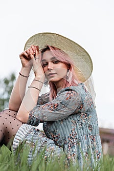 Young lovely woman with pink hair in vintage straw hat in stylish summer blue dress sits on fresh green grass outdoors on nature.