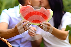 Young, lover`s couple holding a watermelon and kissing, a picnic on the sunflower field