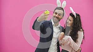 Young lovers couple on the pink background. With hackneyed ears on the head. During this, they make a photo of sephi on