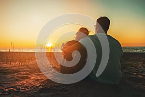 young lovers couple sitting in sand on beach at romantic golden sunset