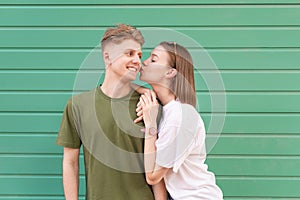 Young lovers are a couple of teens standing on the background of a green wall, the boy smiles, the girl kisses his cheek. Young