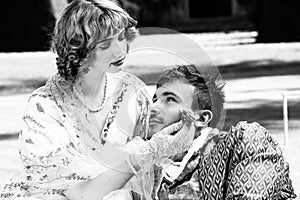 Young lovers dressed in vintage clothing. Man looking into partner`s eyes as she strokes his face with a flower