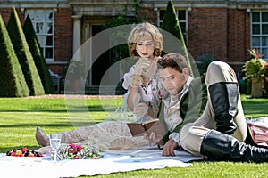 Young lovers dressed in vintage clothing sitting on picnic blanket. Gentleman reading to his lover from a book of poems