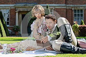Young lovers dressed in vintage clothing sitting on picnic blanket. Gentleman reading to his lover from a book of poems