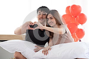 Young lovers are happily making heart symbols by hand to celebrate Valentine`s Day on the bed in their bedroom.