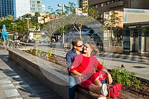 Young loving couple in bright clothes and sunglasse ssitting on the bench in the city area. Embracing, laughing ,relaxing. Happine