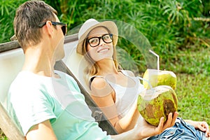 Young loving couple drink coconut while sitting under palms in deck chair lounge resort