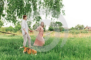 Young loving couple having fun and running on the green grass on the lawn with their beloved domestic dog breed Beagle and a