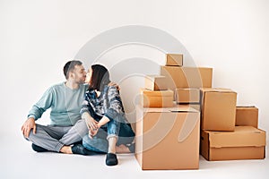 Young loving couple kissing sitting on floor in their new apartment on moving day surrounded by cardboard boxes