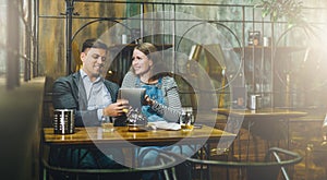 Young loving couple sitting in cafe at table and using tablet computer. Girl and guy use digital gadget, blogging