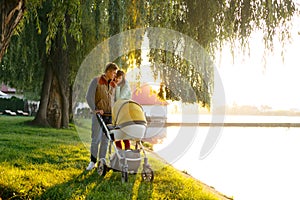 A young loving family walks by the lake with a stroller. Smiling parents couple with baby pram in autumn park. Love, parenthood, f