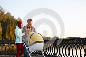 A young loving family walks by the lake with a stroller. Smiling parents couple with baby pram in autumn park. Love, parenthood, f