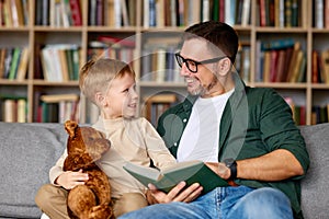 Young loving father reading book to small child son while spending leisure time in living room at home