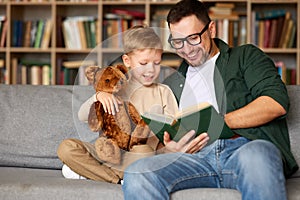 Young loving father reading book to small child son while spending leisure time in living room at home