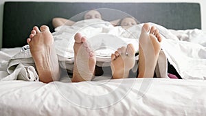 Young loving joyful couple playing and dancing their feet under blanket while wake up in bed in the morning