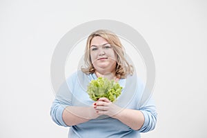 A young lush fat woman in casual blue clothes on a white background holding green lettuce leaves in her hands and mouth.